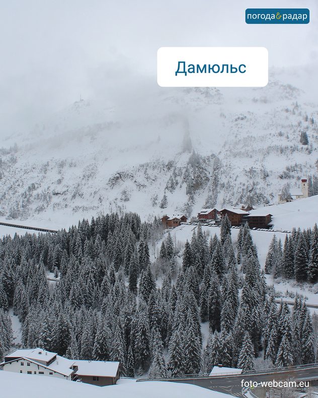 На зображенні з веб-камери видно засніжений гірський пейзаж із гірською дорогою та кількома будинками.