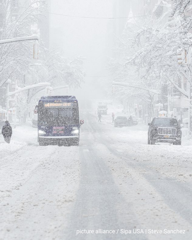 Zasněžená městská ulice na východním pobřeží USA s hustým sněžením. Autobus projíždí vysokými sněhovými závějemi, auta stojí na okraji silnice. Stromy a semafory jsou hustě pokryty sněhem, viditelnost je velmi omezená.