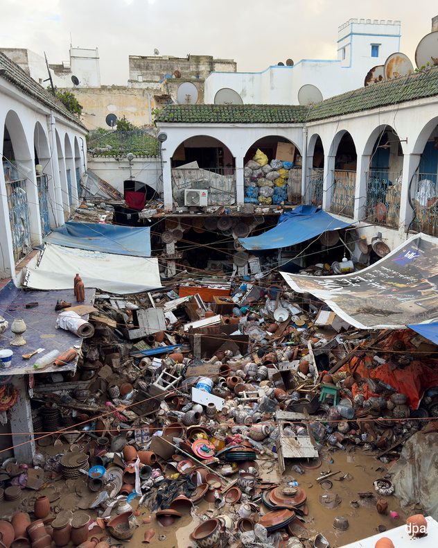 Cour intérieure inondée jonchée de débris et de poteries brisées. Des arcades et des bâches endommagées pendent au-dessus de la zone détruite.
