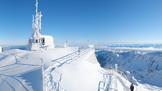 Das Bild zeigt eine verschneite Gipfelstation mit Antenne und Geländer unter blauem Himmel. Im Hintergrund erstrecken sich schneebedeckte Berge über dem Wolkenmeer.