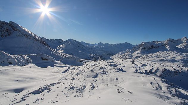 Das Bild zeigt eine sonnige, tief verschneite Berglandschaft unter strahlend blauem Himmel. Die Sonne steht tief und lässt den Schnee hell glitzern. (c) foto-webcam.eu