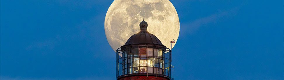 Vollmond steht direkt hinter der Laterne eines rot gemauerten Leuchtturms und erscheint vor dem blauen Abendhimmel wie eine leuchtende Scheibe. (c) Klaus Haase