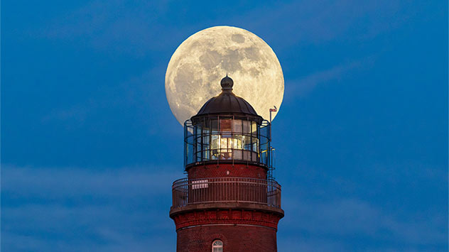 Vollmond steht direkt hinter der Laterne eines rot gemauerten Leuchtturms und erscheint vor dem blauen Abendhimmel wie eine leuchtende Scheibe.