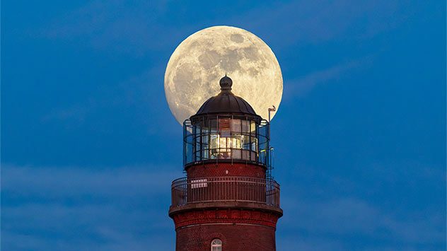 Vollmond steht direkt hinter der Laterne eines rot gemauerten Leuchtturms und erscheint vor dem blauen Abendhimmel wie eine leuchtende Scheibe. (c) Klaus Haase