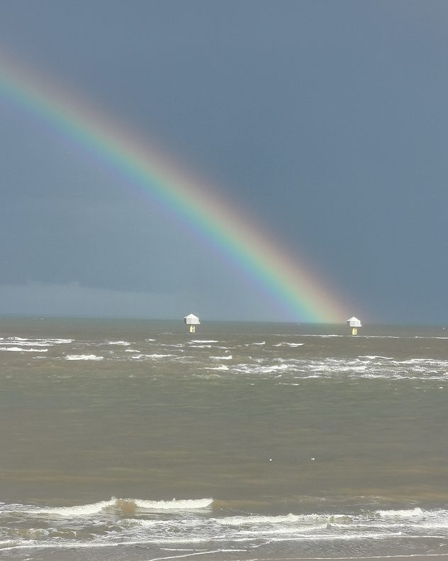 Bright rainbow descending into a choppy gray sea beneath dark storm clouds, with whitecaps on the water and two offshore navigation beacons near the horizon.