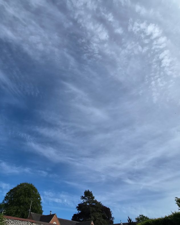 Wide sky filled with rippled altocumulus cloud layers and wispy streaks above rooftops and trees, creating a textured mackerel-sky pattern against a blue backdrop.