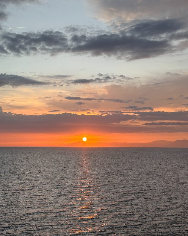 Sun setting over a calm sea with orange and pink light reflecting across the water, silhouetted distant mountains on the horizon, and layered clouds overhead.