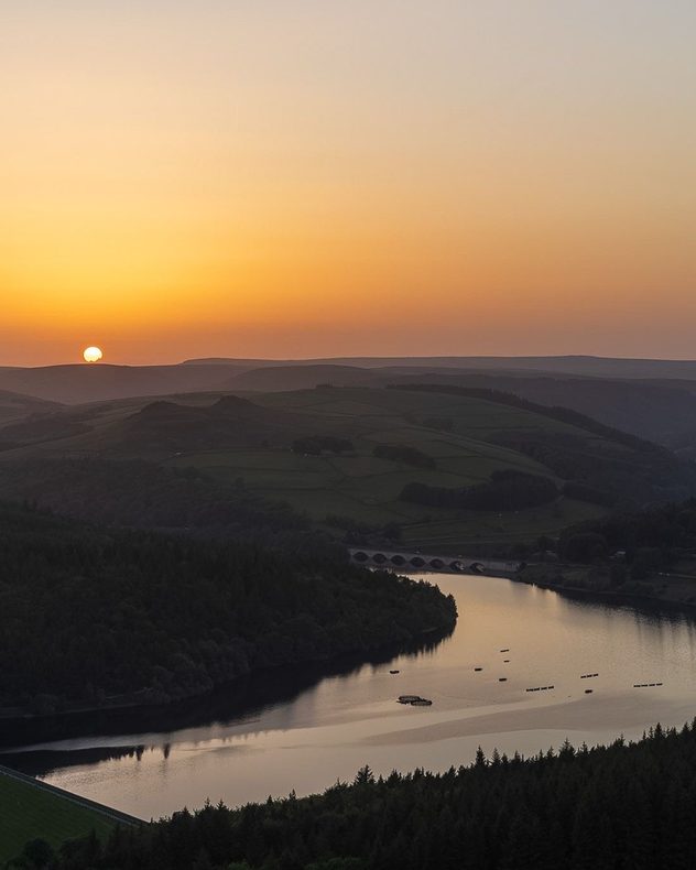 Sunset over a calm reservoir winding through rolling hills and forested valleys, with golden-orange skies, dark silhouettes, and reflections on the water below.