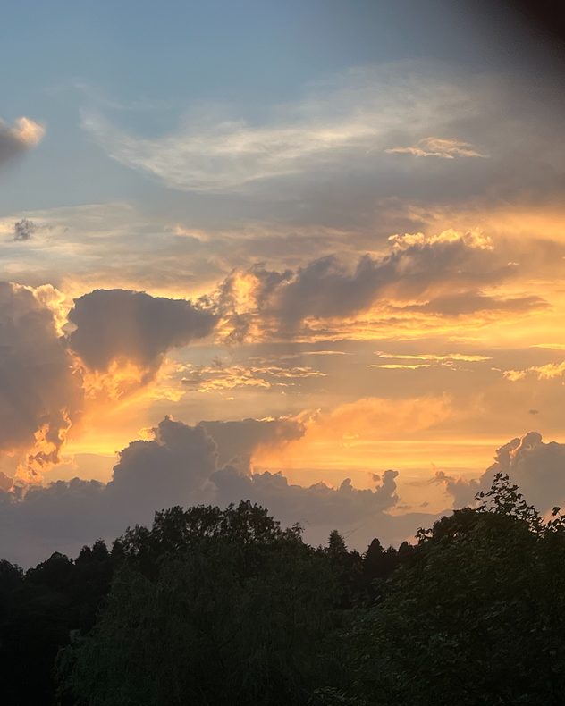 Towering dark clouds illuminated by golden sunset light above a tree-lined horizon, with glowing orange and yellow cloud edges against a fading blue sky.
