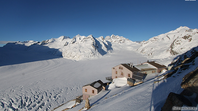 Blick auf den verschneiten Aletschgletscher mit umliegenden Gipfeln und einer Berghütte im Vordergrund bei klarem Himmel.