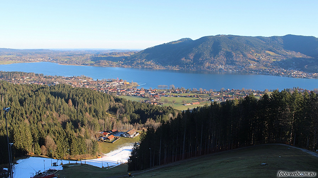 Blick auf den Tegernsee mit umliegenden Wäldern und Ortschaften, im Vordergrund ein schneearmes Skigebiet unter klarem Himmel.