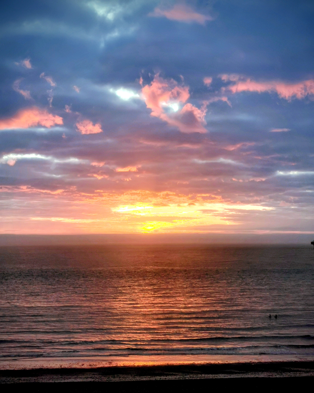 Golden sunset over calm ocean with layered blue and pink clouds above, warm ight reflecting across rippling water, and gentle waves along the shoreline.