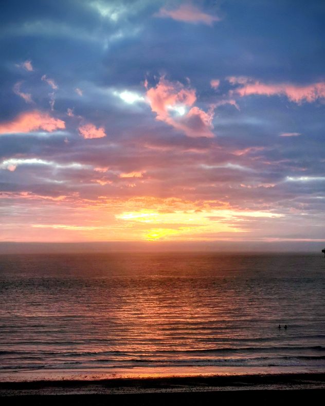 Golden sunset over calm ocean with layered blue and pink clouds above, warm ight reflecting across rippling water, and gentle waves along the shoreline.
