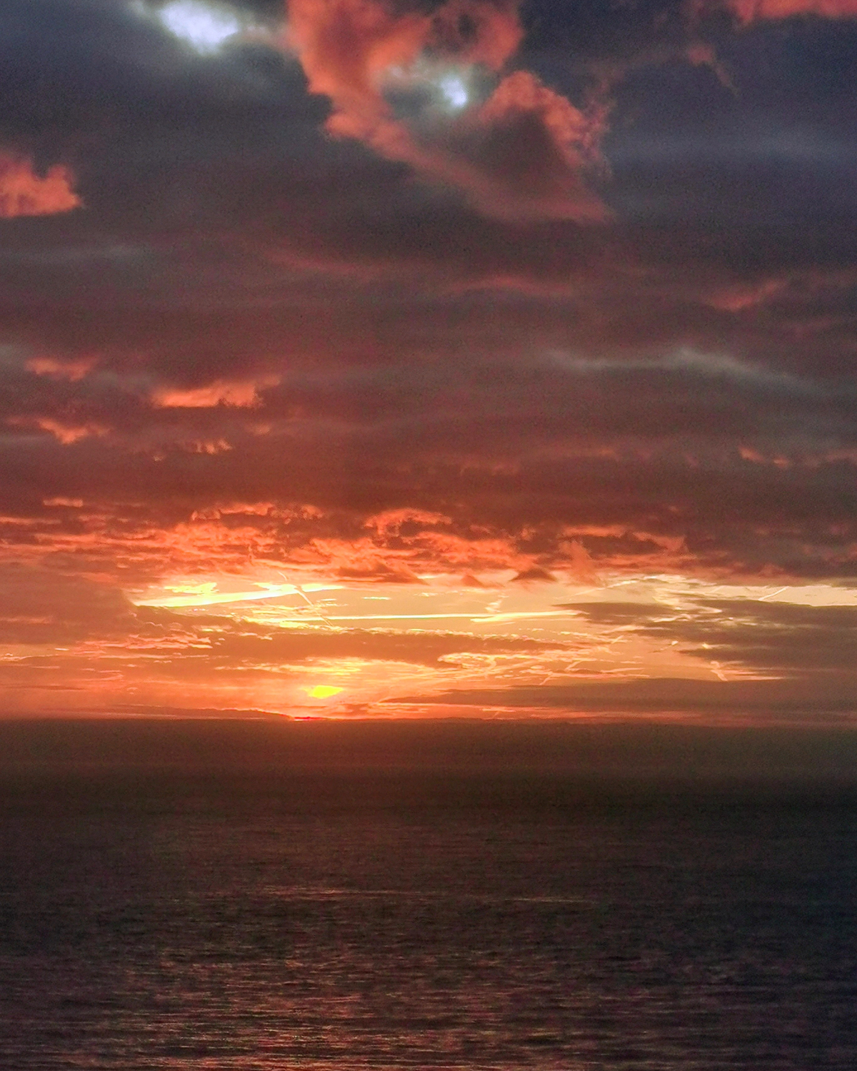 Fiery red and orange sunrise over the sea, with dark layered clouds glowing above the horizon and warm reflections shimmering across the water.
