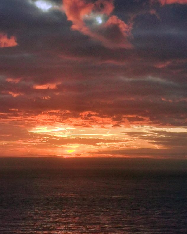 Fiery red and orange sunrise over the sea, with dark layered clouds glowing above the horizon and warm reflections shimmering across the water.