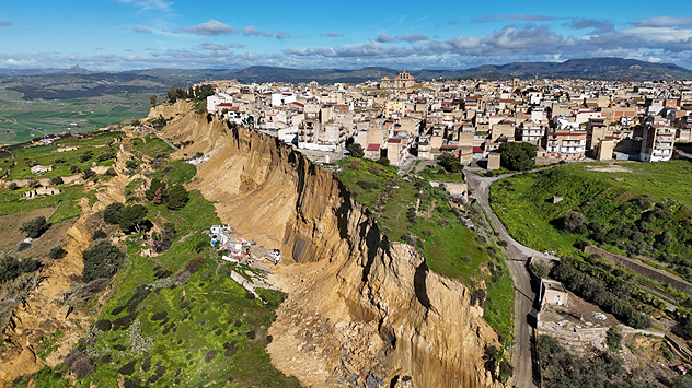 Ciudad densamente urbanizada al borde de un acantilado rocoso escarpado y erosionado. En primer plano, laderas verdes y profundos desprendimientos.