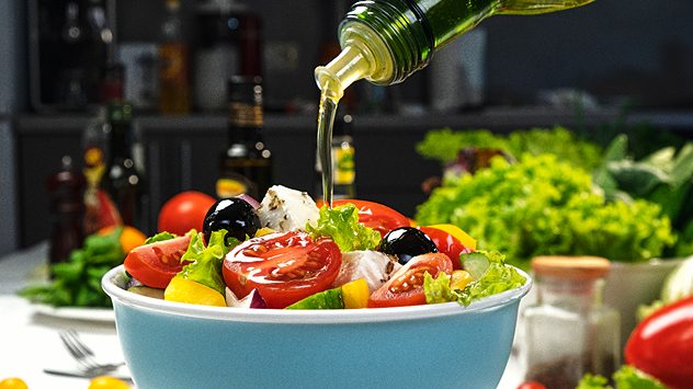 In eine bunte SchÃ¼ssel mit frischem Salat aus Tomaten, Gurken, Paprika, Oliven und Feta wird OlivenÃ¶l gegossen â im Hintergrund frisches GemÃ¼se und Flaschen auf einem KÃ¼chentisch. (c) Spalnic/Shutterstock.com