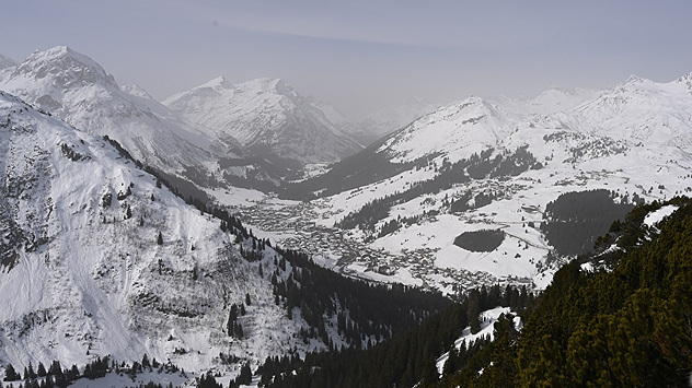 Verschneites Alpendorf in einem Tal, umgeben von hohen Bergen. Winterlandschaft mit dichtem Schnee und trübem Himmel.