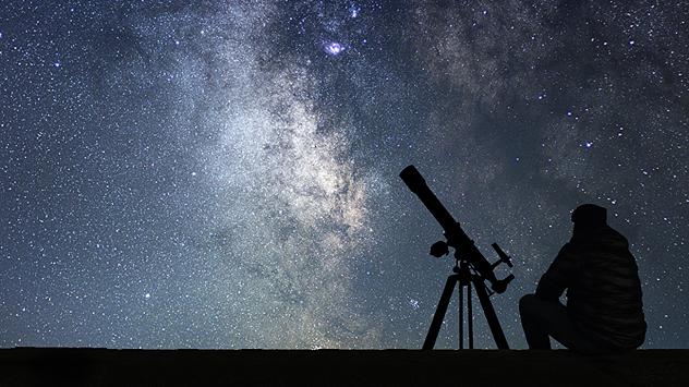 Silhouette of a person sitting beside a telescope on a tripod under a clear night sky filled with dense stars and the bright band of the Milky Way.