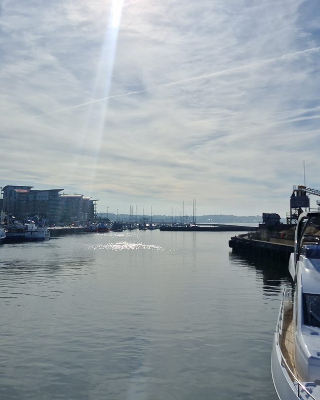 Calm marina under bright sunshine with boats, still water, and thin cirrus and cirrostratus clouds crossing a pale blue sky, while sunlight reflects off the harbour surface.