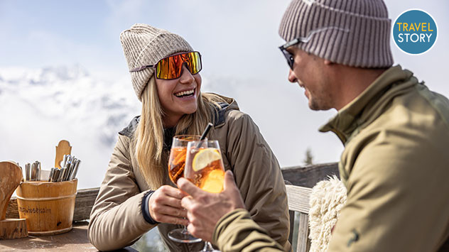 Ein Paar stößt auf einer sonnigen Bergterrasse mit Aperol Spritz an, im Hintergrund schneebedeckte Berge.