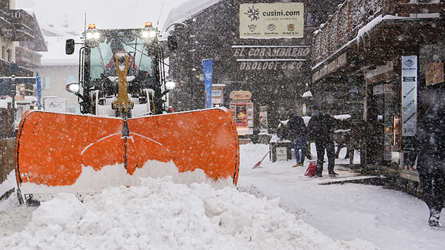 The picture shows a mountain village during heavy snowfall. A snowplough clears the road of snow.
