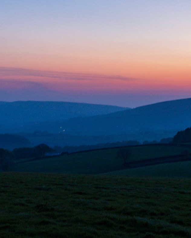 Rolling hills and dark grassy fields at dawn beneath a pink and purple sunset sky, with layered mountain silhouettes fading into the distance.