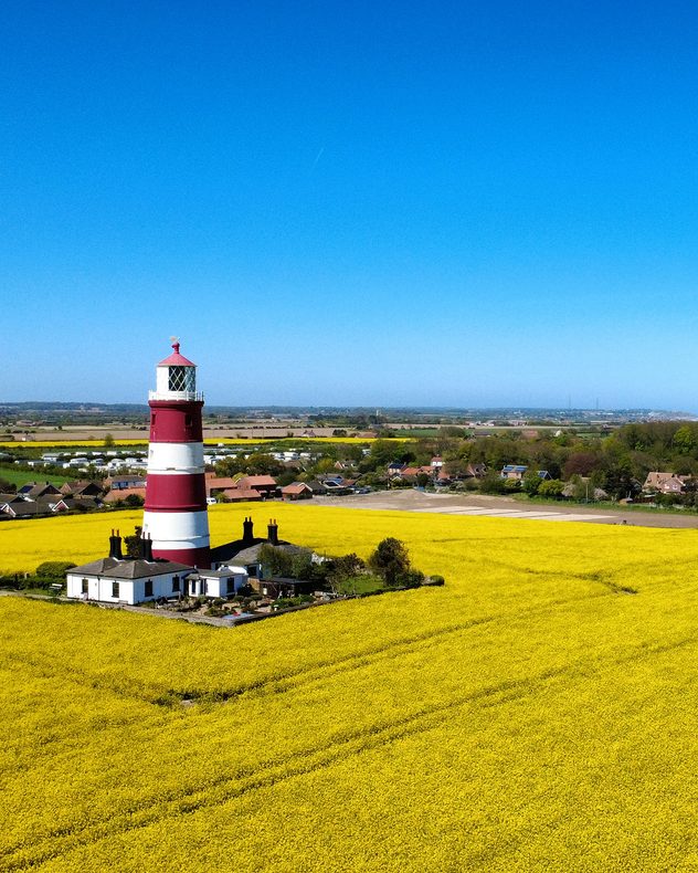 Red and white striped lighthouse surrounded by bright yellow fields under a clear blue sky, with rural houses and farmland in the distance.