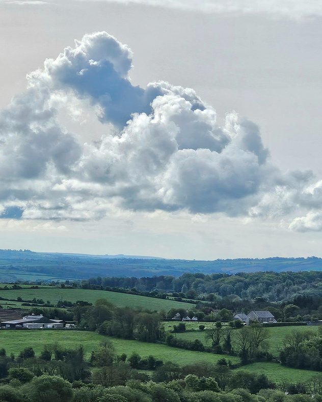 Large towering cumulus clouds above rolling green countryside with scattered trees, farm buildings, and soft sunlight breaking through the grey sky.