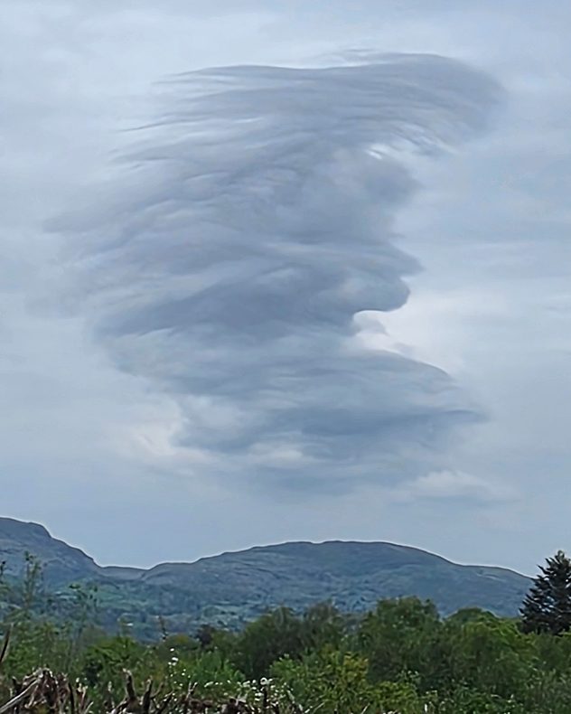 Large layered lenticular cloud with swirling textures above low mountains and green trees under a gray overcast sky.
