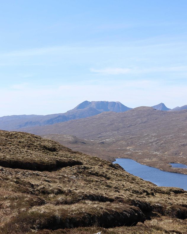 Rocky mountain landscape with narrow blue loch winding through barren hills beneath a pale blue sky and distant rugged peaks.