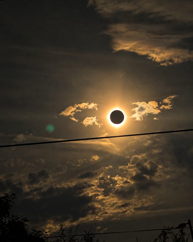 Total solar eclipse with the Moon covering the Sun and a bright golden corona glowing through dramatic dark clouds, with silhouetted wires crossing the sky.