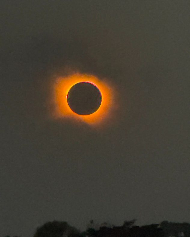 Total solar eclipse with the Moon completely covering the Sun, revealing a glowing orange corona and a bright prominence along the left edge against a dark sky.