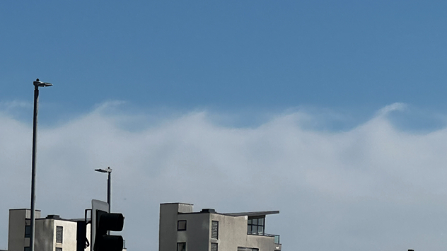 Kelvin-Helmholtz wave clouds forming rolling crest shapes above modern buildings under a clear blue sky, with smooth white cloud bands resembling breaking ocean waves.