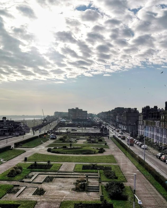 Elevated view of a coastal seafront with a formal park and pathways in the foreground, terraced houses lining the road to the right, light traffic, birds in the sky, and broken cloud with sunlight over the sea.