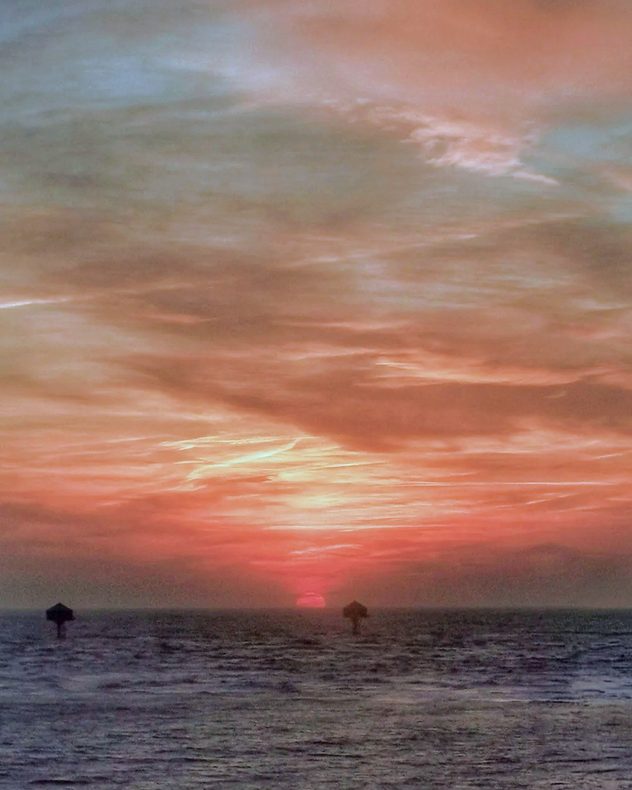 Sun setting over the sea with layered clouds glowing red, orange, and pale blue, dark water in the foreground, and small navigation markers silhouetted near the horizon.