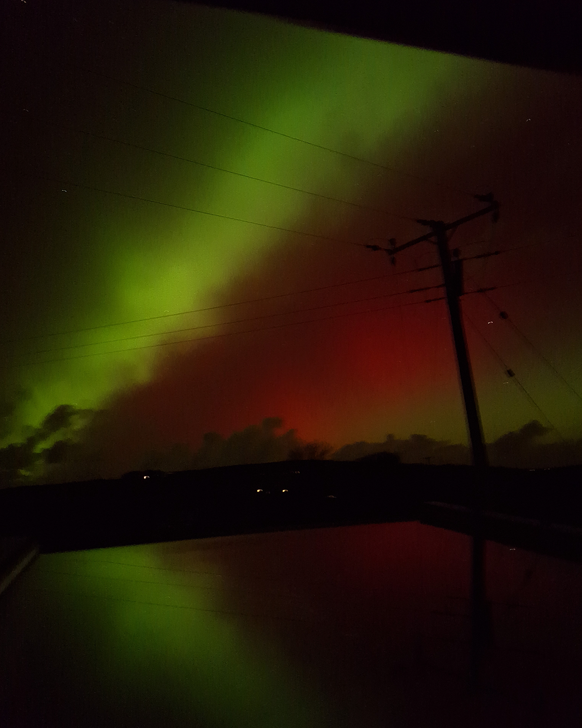 Green aurora bands stretching across the night sky with a red glow beneath, crossed by power lines and a silhouetted utility pole above a dark horizon.