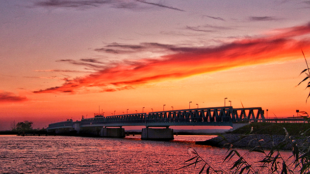 © Anke Hanusik Leuchtend roter Himmel bei Sonnenuntergang. Im Vordergrund fließt ein Fluss, im Hintergrund erstreckt sich eine Brücke mit Laternenmasten.