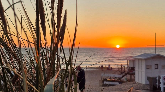 © Sylter Sekunden by Uwe Schmale Sonnenuntergang am Meer mit Blick durch Strandgras. Am Strand stehen weiße Hütten und einige Menschen.