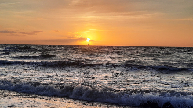 © Ralf Kammer via WetterMelder Deutschland Die Sonne geht direkt über dem Meer unter. Der Himmel ist orange gefärbt, Wellen schlagen schäumend ans Ufer.