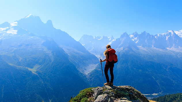 Eine Wanderin mit rotem Rucksack steht auf einem felsigen Aussichtspunkt und blickt auf ein weites Tal und schneebedeckte Berggipfel unter blauem Himmel.