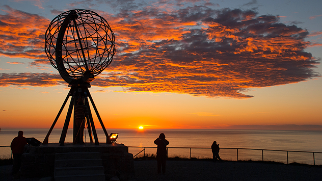 Sonnenuntergang über dem Meer am Nordkap in Norwegen. Im Vordergrund die bekannte Globus-Skulptur, daneben Menschen, die den Anblick fotografieren. Der Himmel ist in Orange- und Rottönen gefärbt.