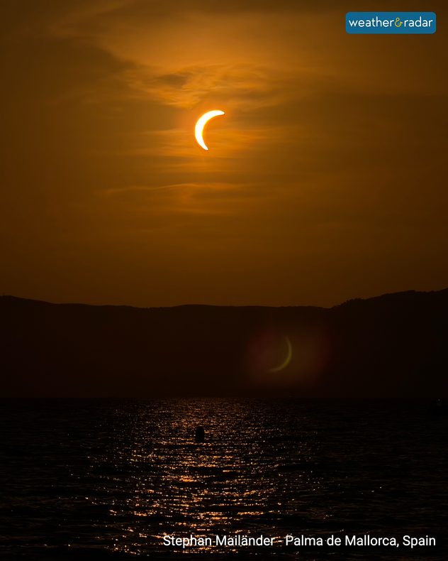 Crescent Sun during a partial solar eclipse above dark hills and shimmering sea at sunset in Palma de Mallorca, Spain, beneath thin orange clouds.