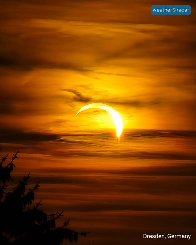 Partial solar eclipse glowing through layered orange clouds at sunset in Dresden, Germany, with the Sun reduced to a bright curved crescent.
