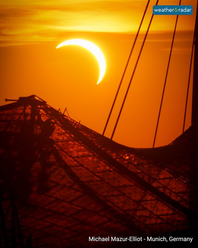 Bright crescent Sun during a partial solar eclipse in an orange sunset sky above silhouetted structural cables and framework in Munich, Germany.