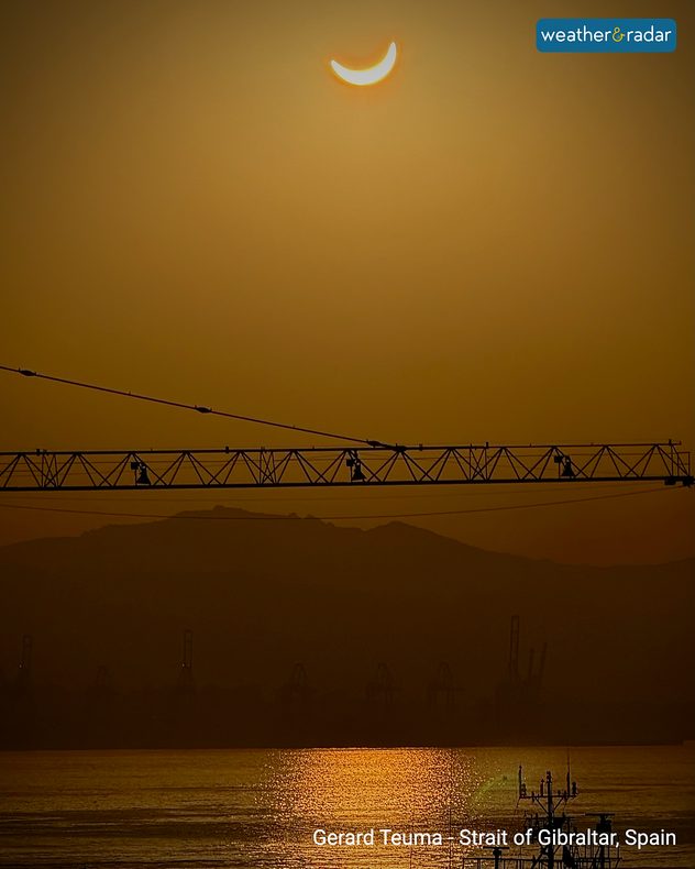 Crescent Sun during a partial solar eclipse above Gibraltar Bay, with silhouetted cranes, distant hills and golden light reflecting across the water.