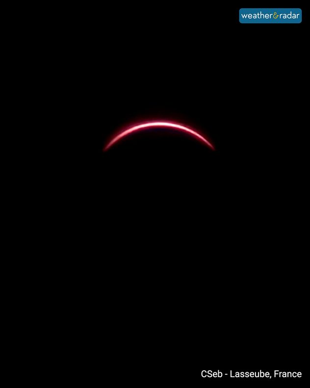 Thin pink-red arc of the Sun visible during a deep partial solar eclipse against a black sky, photographed in Lasseube, France.