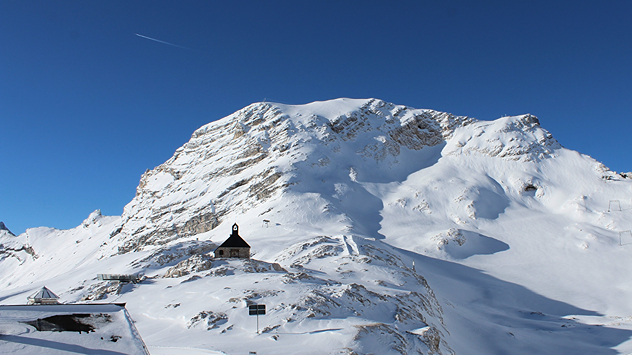 Das Bild zeigt eine schneebedeckte Berglandschaft mit einer kleinen Kapelle im Vordergrund. Der Himmel ist klar und tiefblau, die Sonne beleuchtet die weißen Hänge.