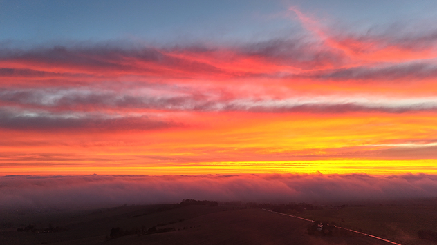 Leuchtender Sonnenaufgang über nebelbedeckten Feldern. Rote und orangefarbene Wolken dominieren den Horizont.