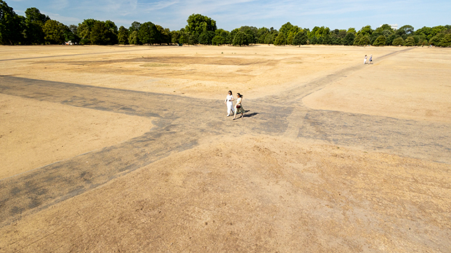 Aerial view of a large park with dry yellow-brown grass, intersecting footpaths, and a few people walking across the parched landscape beneath clear summer skies.